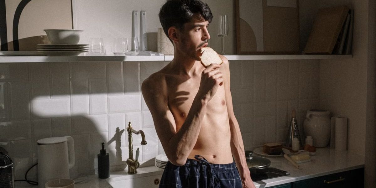 Shirtless young man bites toast in bright kitchen morning scene.