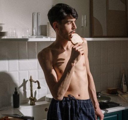 Shirtless young man bites toast in bright kitchen morning scene.