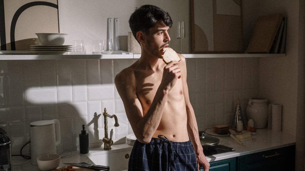 Shirtless young man bites toast in bright kitchen morning scene.